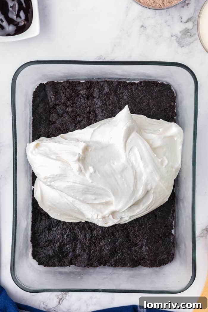 Cream cheese mixture being spread over the Oreo crumb crust in a glass dish.
