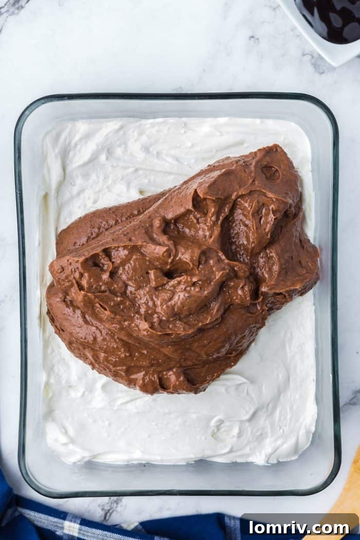 Chocolate pudding being spread over the cream cheese mixture in a glass dish.