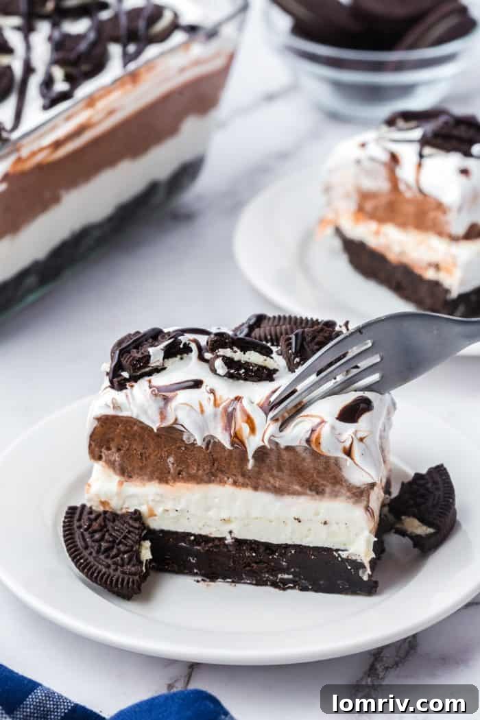 Two plates with slices of no bake Oreo layered dessert with the glass dish with the dessert in it in the background. One slice of dessert has a fork going into it.