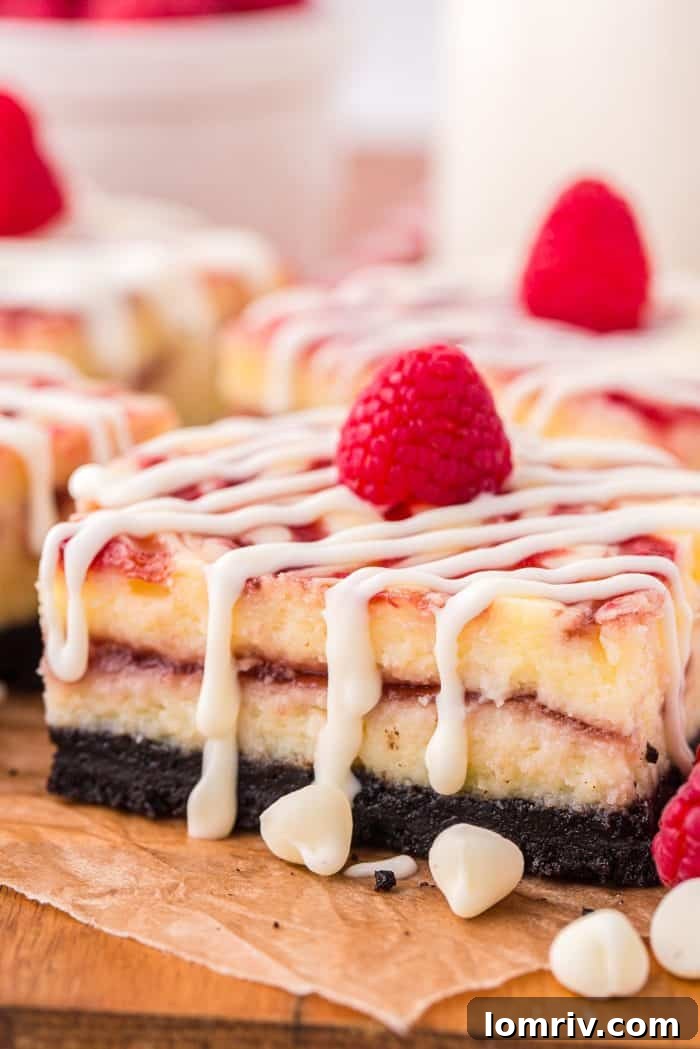A closer view of four finished white chocolate raspberry cheesecake bars, garnished with fresh raspberries, on parchment paper with scattered white chocolate chips.