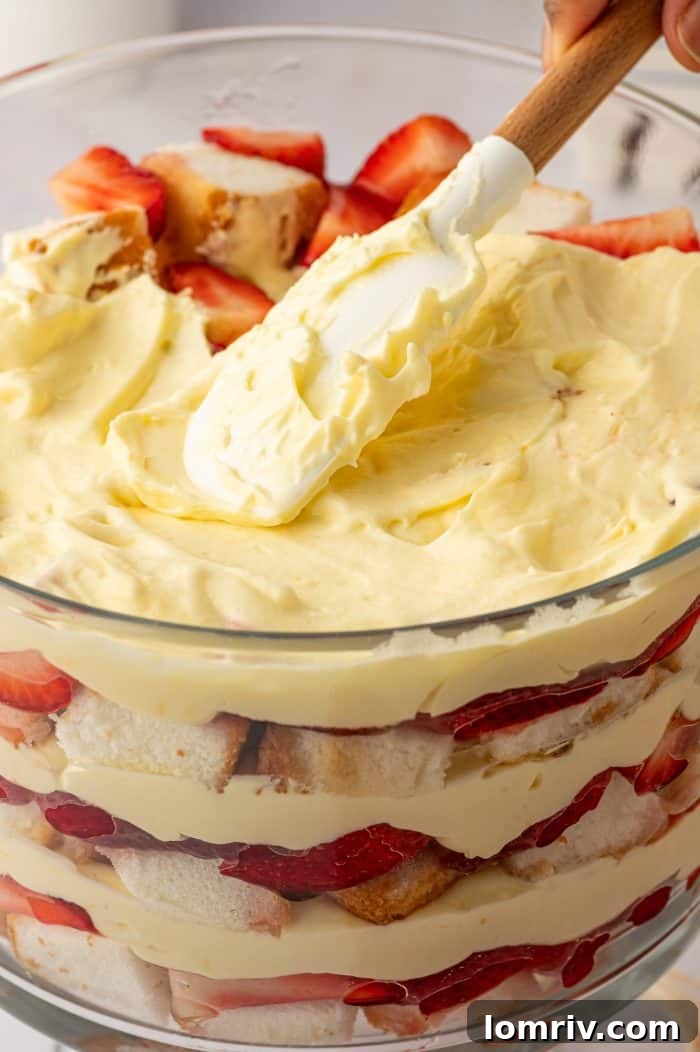 A close-up shot of the trifle during assembly, showing a knife carefully spreading a smooth layer of pudding over the fruit and cake.