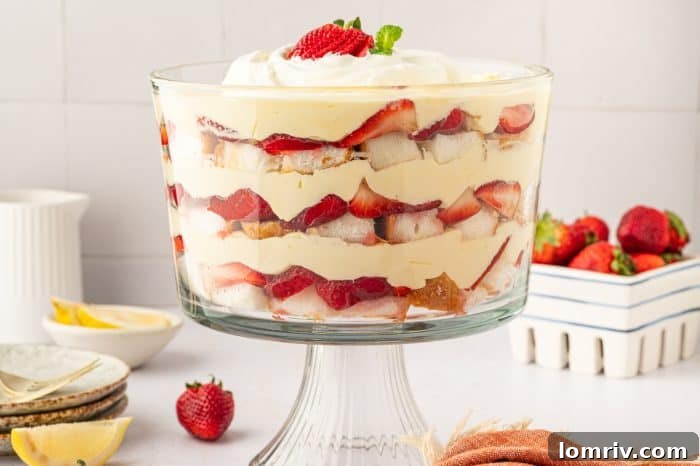 The full strawberry trifle beautifully displayed in a large glass stand, with fresh strawberries artfully arranged in the soft background.
