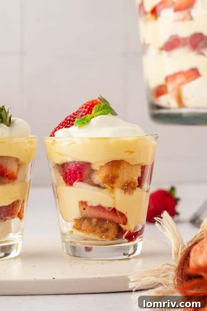 Two individual portions of strawberry trifle in glass cups, set against a backdrop of a larger trifle bowl filled with the same dessert.