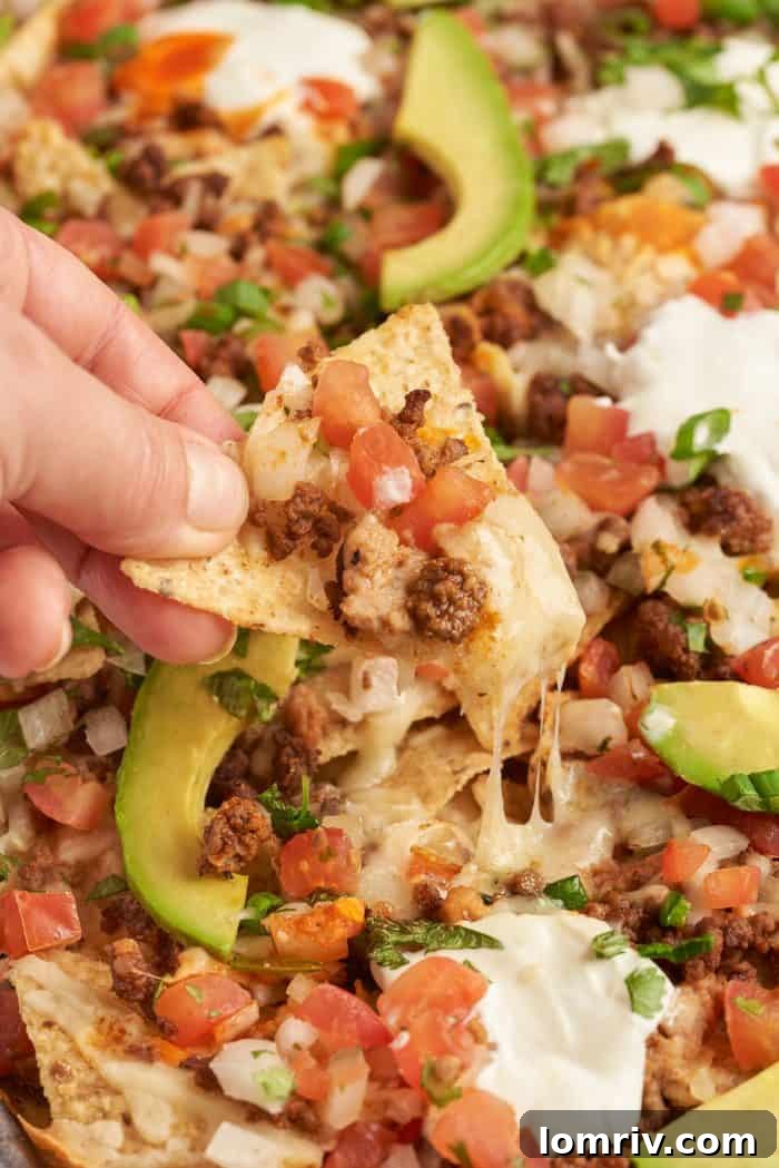 Baked sheet pan nachos with pico de gallo and sliced avocado. A hand is picking up a tortilla chip with melted cheese and toppings.