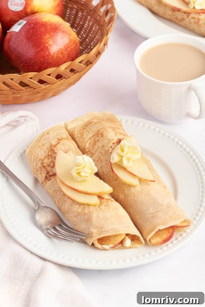 A white plate with two apple crepes that are filled with a cooked apple filling and garnished with sliced apples.