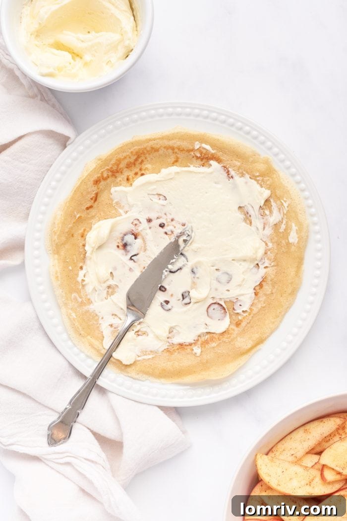 A white plate with a crepe and mascarpone being spread on it.