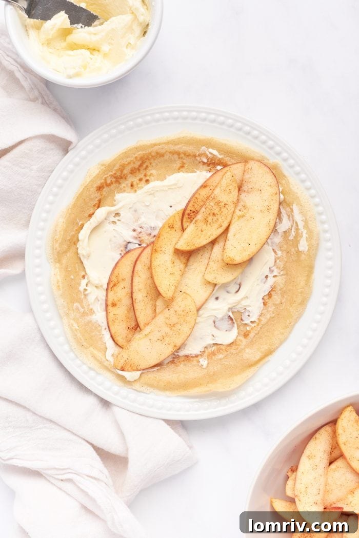 A white plate with a crepe, mascarpone, and cooked apple slices.