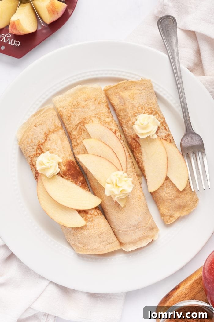 A white plate with 3 rolled crepes filled with cooked apples, and garnished with apple slices.