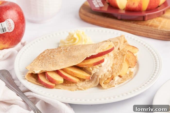 A white plate with two apple crepes that are filled with a cooked apple filling and garnished with sliced apples.
