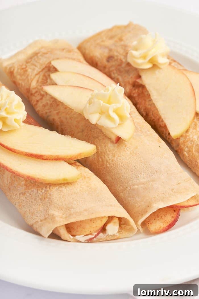 A white plate with three apple crepes that are filled with a cooked apple filling and garnished with sliced apples.