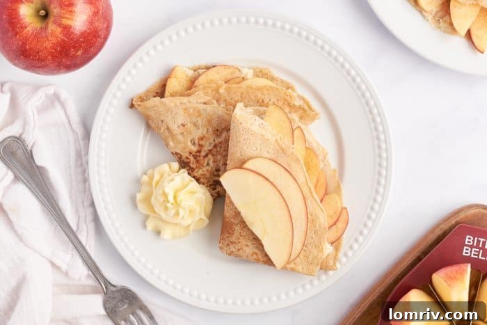 A white plate with two apple crepes that are filled with a cooked apple filling and garnished with sliced apples.