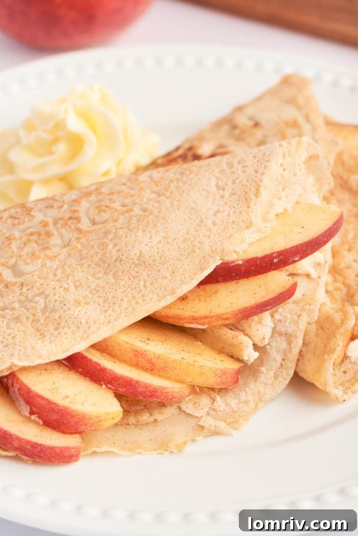 A white plate with a crepe filled with cooked apples, and garnished with apple slices.