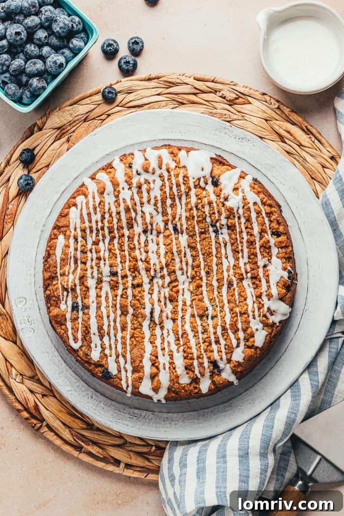 A stunning whole blueberry sour cream coffee cake, generously drizzled with a sweet vanilla glaze, presented on a large white serving plate.