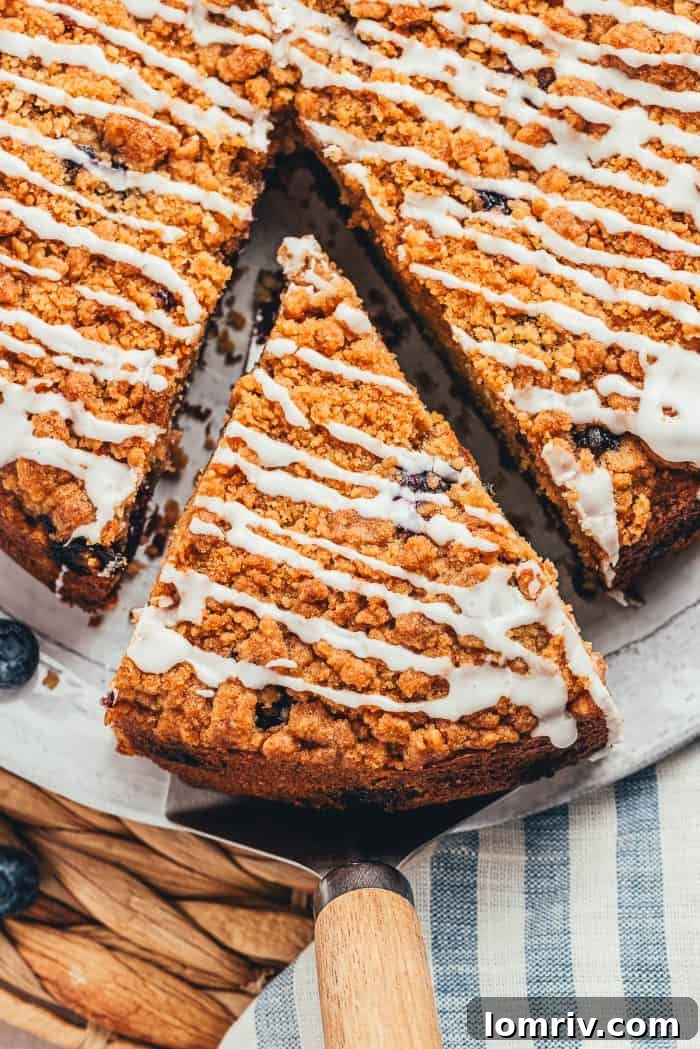 A hand gently removing a perfect slice of blueberry sour cream coffee cake from the whole cake, highlighting its rich layers and crumbly top.