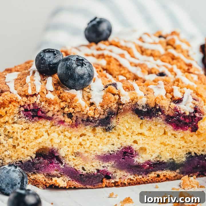 A slice of blueberry sour cream coffee cake on a white plate, showcasing its moist crumb, juicy blueberries, and delicate glaze.