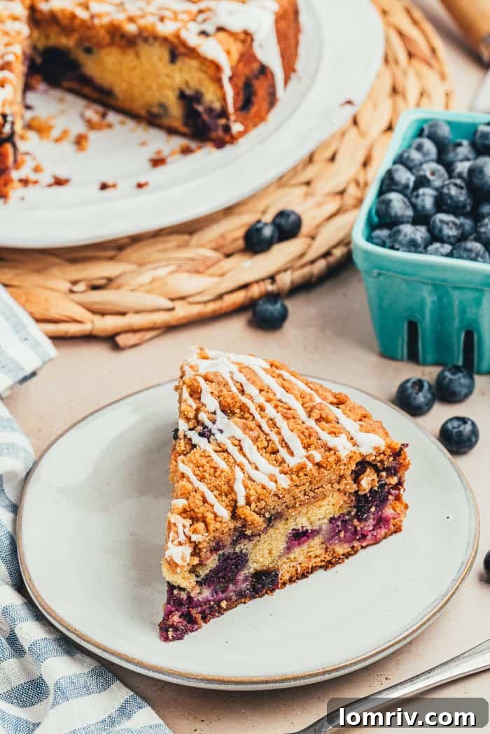 A close-up shot of a single slice of blueberry sour cream coffee cake on a white plate, showcasing the delicate glaze and tender, fruit-filled interior.