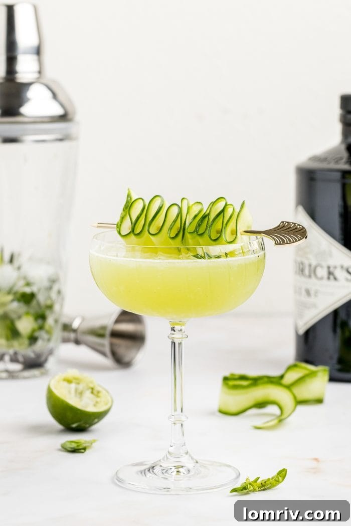 A beautifully garnished Cucumber Basil Gimlet with a delicate cucumber ribbon and fresh basil sprig, set against a blurred green background.