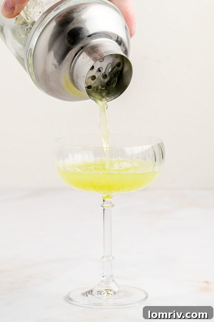 A smooth, vibrant Cucumber Basil Gimlet being expertly poured from a shaker into a coupe glass, ready for enjoyment.