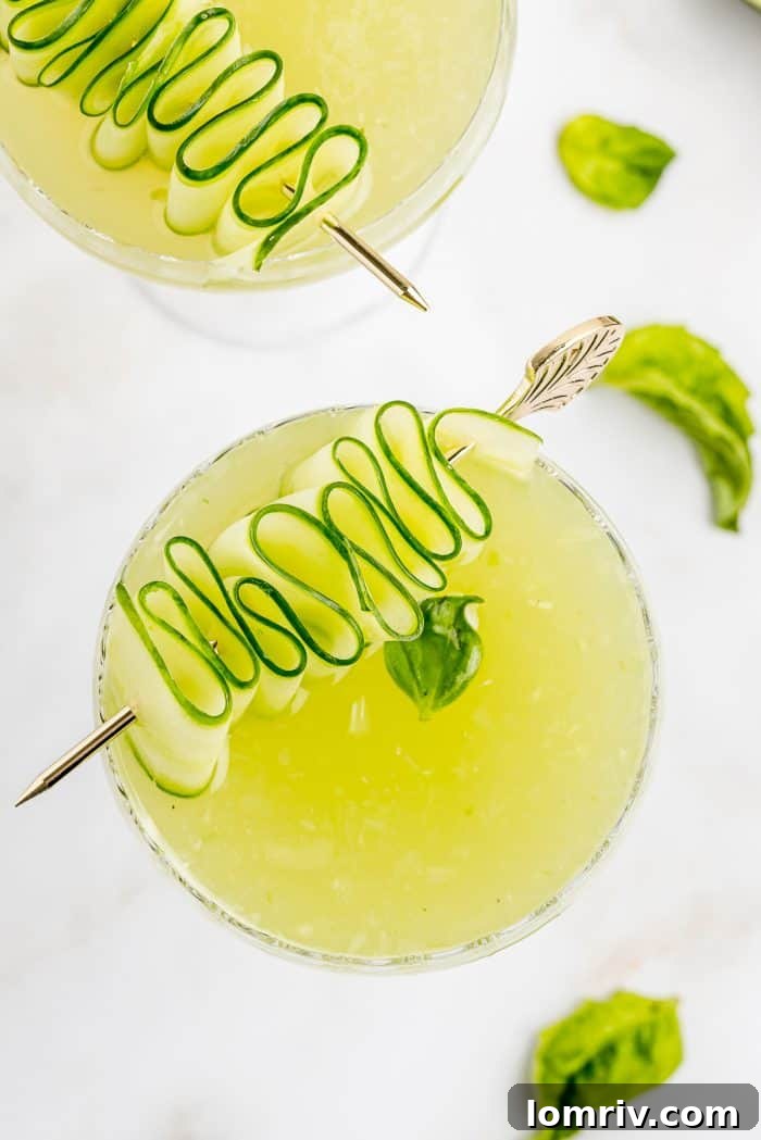 A stunning close-up of a Cucumber Basil Gimlet, featuring a perfectly coiled cucumber ribbon garnish and a fresh basil leaf, highlighting its vibrant green color.