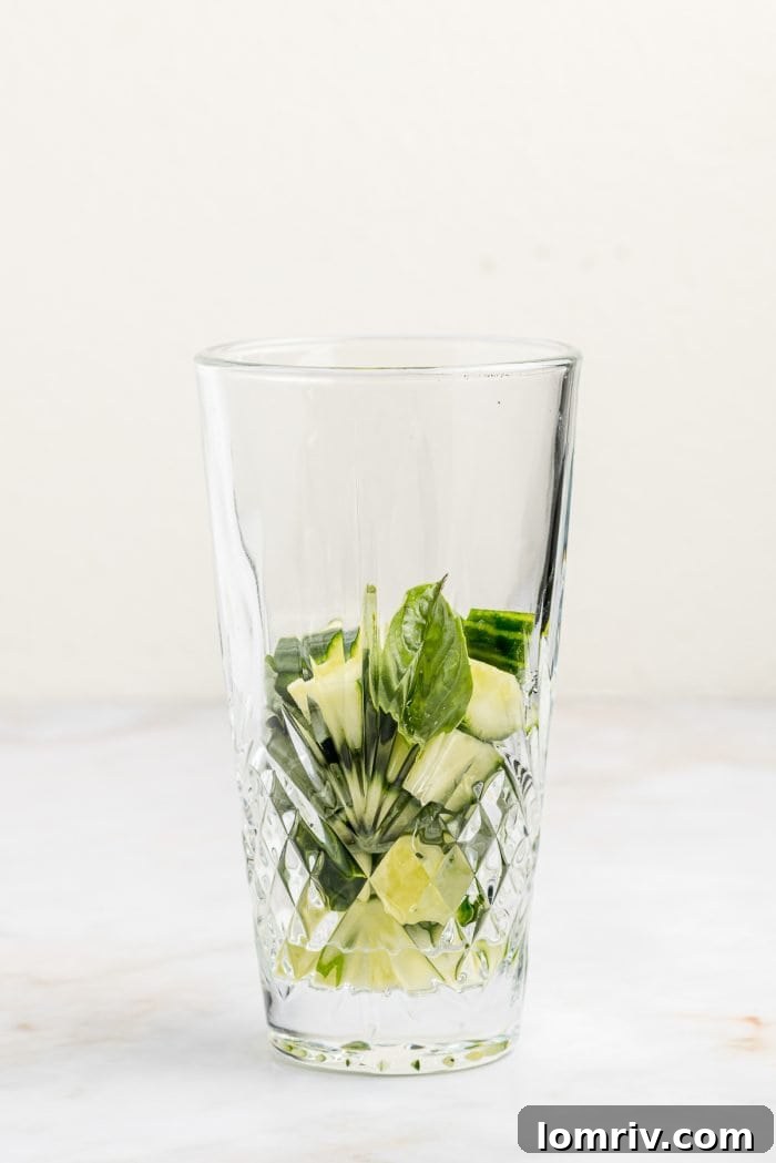 A clear cocktail shaker containing diced cucumber and fresh basil leaves, ready for muddling to release their aromatic flavors.