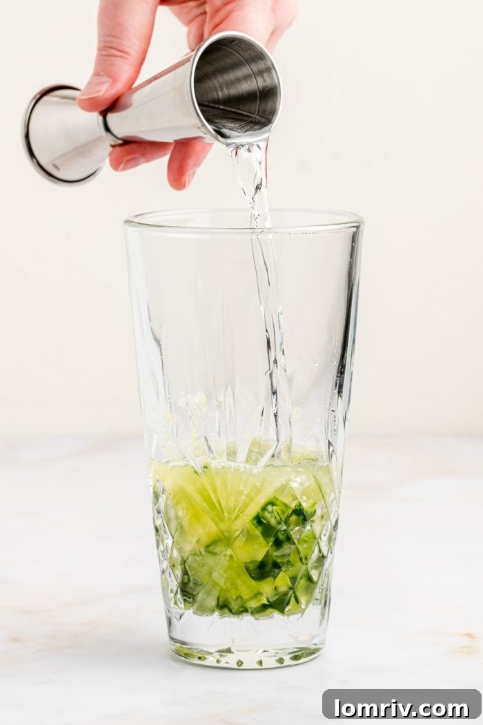 Pouring gin into a cocktail shaker filled with muddled cucumber, basil, and lime juice, preparing for the perfect shake.