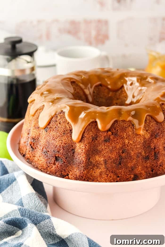 A stunning Caramel Apple Bundt Cake, generously drizzled with rich caramel sauce, displayed on an elegant white cake stand, inviting fall festivities.