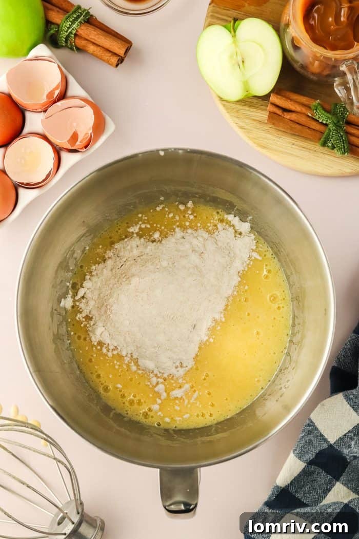 The rich, thick cake batter, a harmonious blend of wet and dry ingredients, fills a metal mixing bowl, showcasing its readiness for the apples.