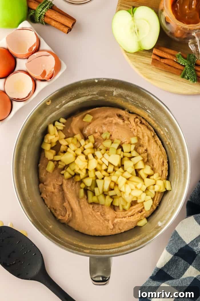 Finely diced apples, now infused with flavor, are gently folded into the robust cake batter within a metal mixing bowl, promising a burst of fruit in every bite.