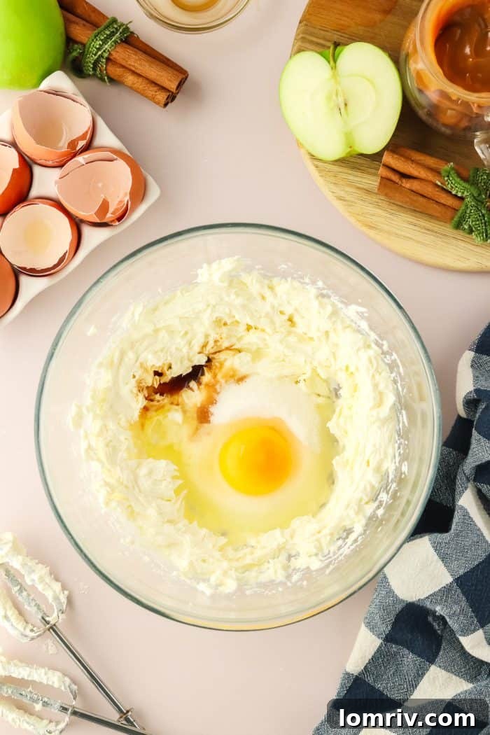 Cream cheese, an egg, and vanilla extract are being whipped together in a clear glass mixing bowl, forming the luscious, smooth cheesecake swirl.
