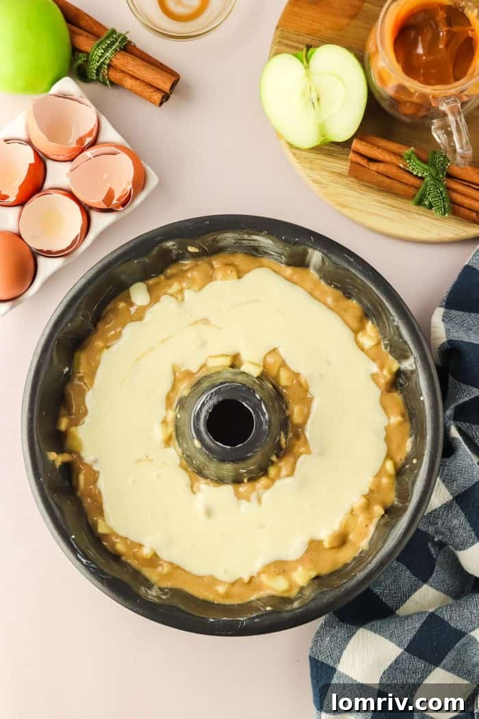 The first layer of apple cake batter is evenly spread in a Bundt pan, crowned with a smooth, inviting layer of cream cheese mixture, ready for the final batter.