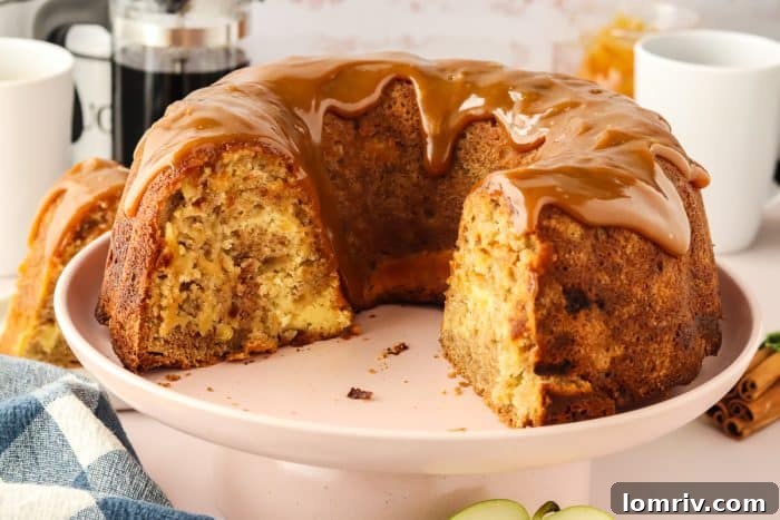 A glorious Caramel Apple Bundt Cake, missing a few slices, sits elegantly on a white cake plate, its rich caramel drizzle a testament to its autumnal indulgence.