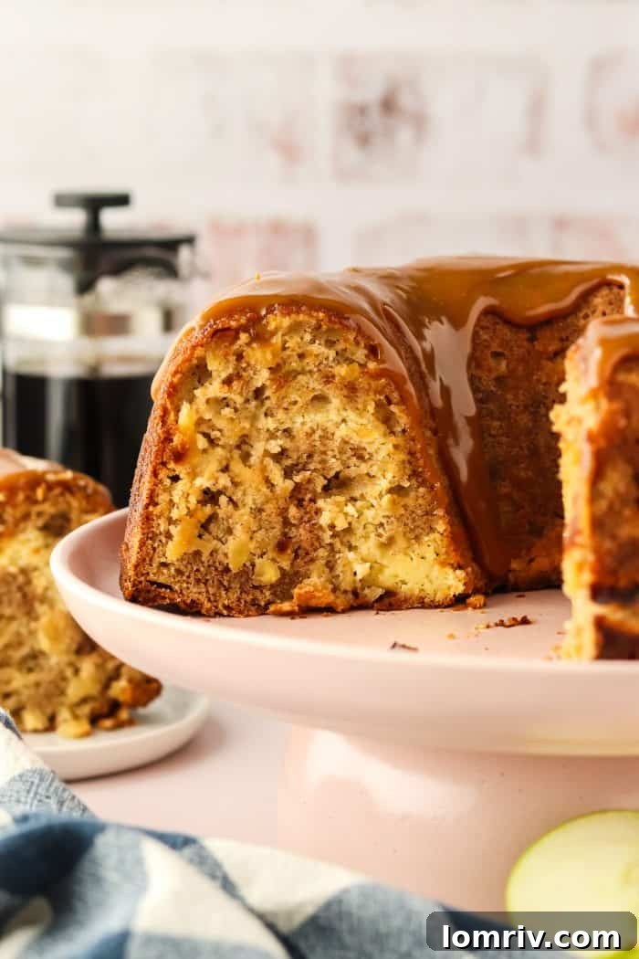 The stunning Caramel Apple Bundt Cake, with several slices already removed, emphasizing its generous caramel drizzle and inviting presentation on a pristine white cake plate.