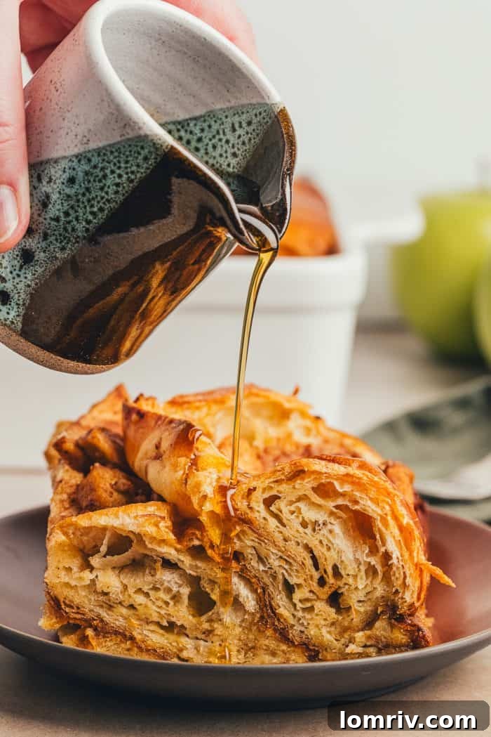 A slice of apple french toast bake on a brown colored plate with a bottle of maple syrup being poured over it.