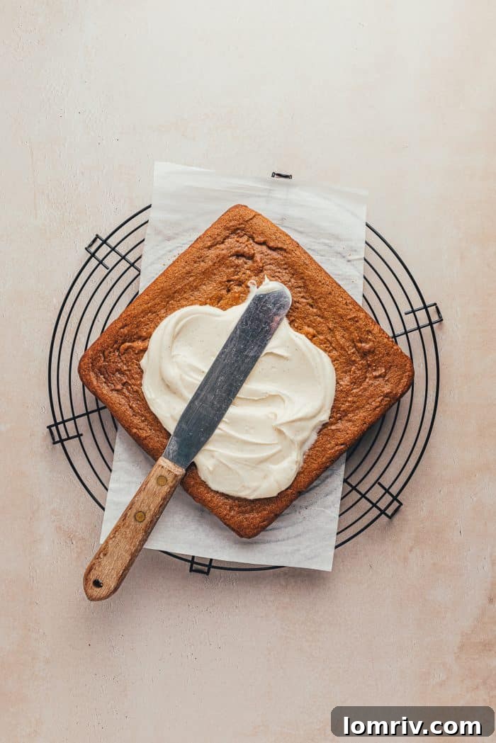 A spatula smoothly spreading creamy white frosting over the top of baked gingerbread bars in a pan.