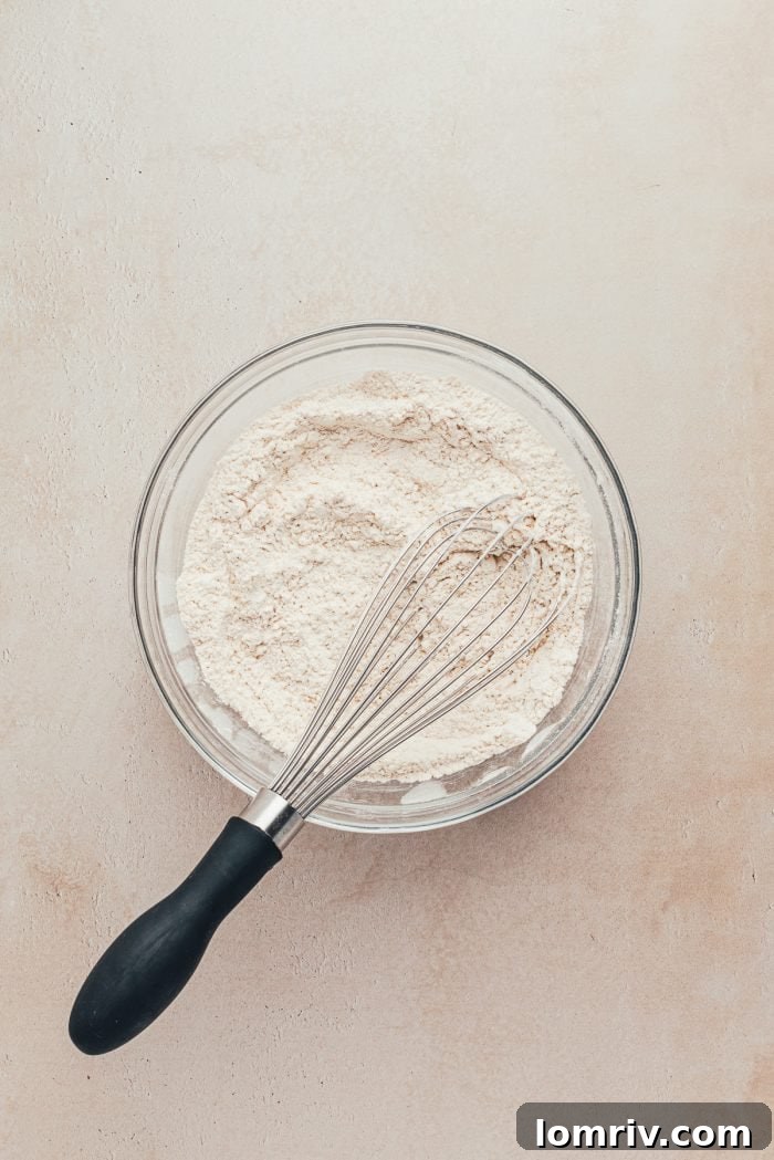 A glass bowl holds the dry ingredients for gingerbread bars: flour, baking soda, salt, and spices, with a whisk resting inside.