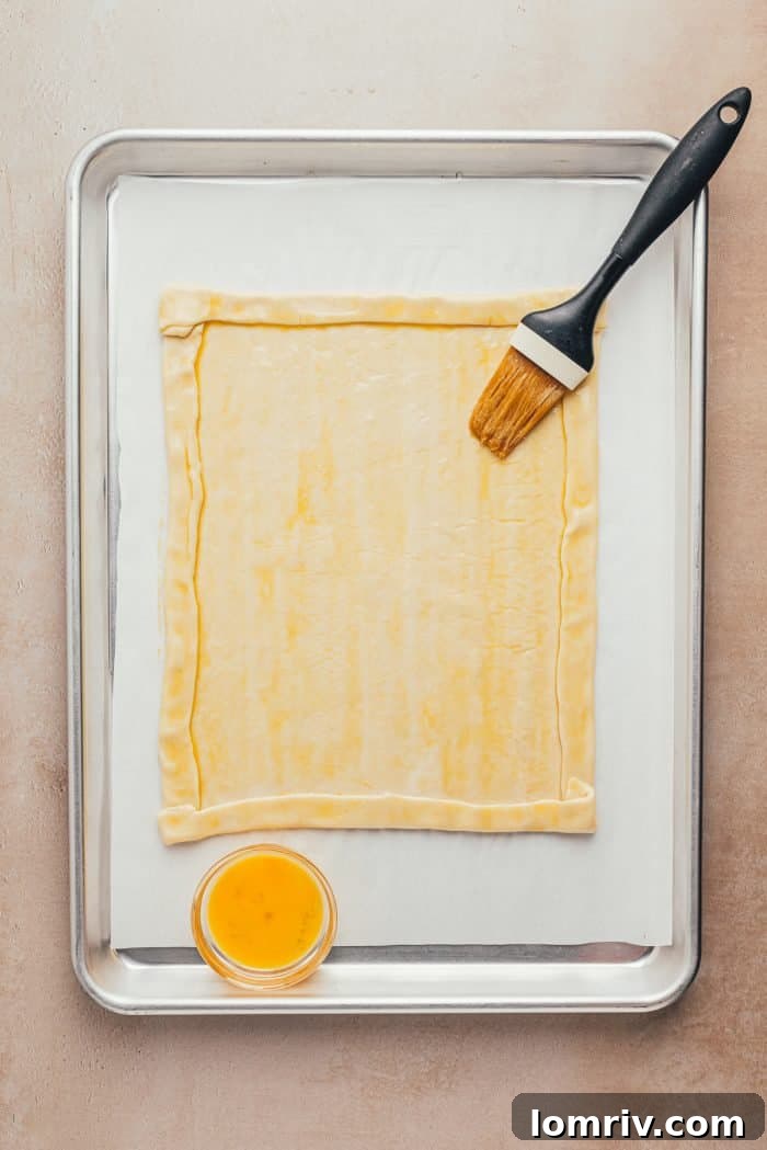 Puff pastry on a baking sheet with a neatly formed crust, being brushed with an egg wash using a pastry brush.