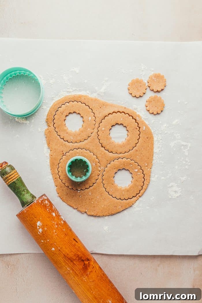 Parchment paper with rolled-out Linzer dough, adorned with cookie cutters for both the main shape and the smaller center cutout, alongside a rolling pin.