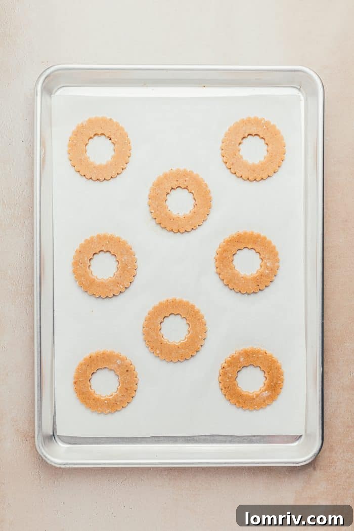 A baking tray with parchment paper, carefully arranged with unbaked Linzer cookie tops, each featuring a festive center cutout.