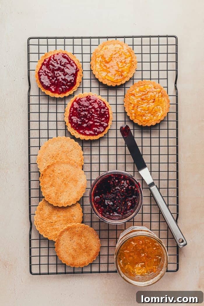 Baked Linzer cookie bottoms on a cooling rack, with jam being spread on them, next to a knife and two bowls of jam.
