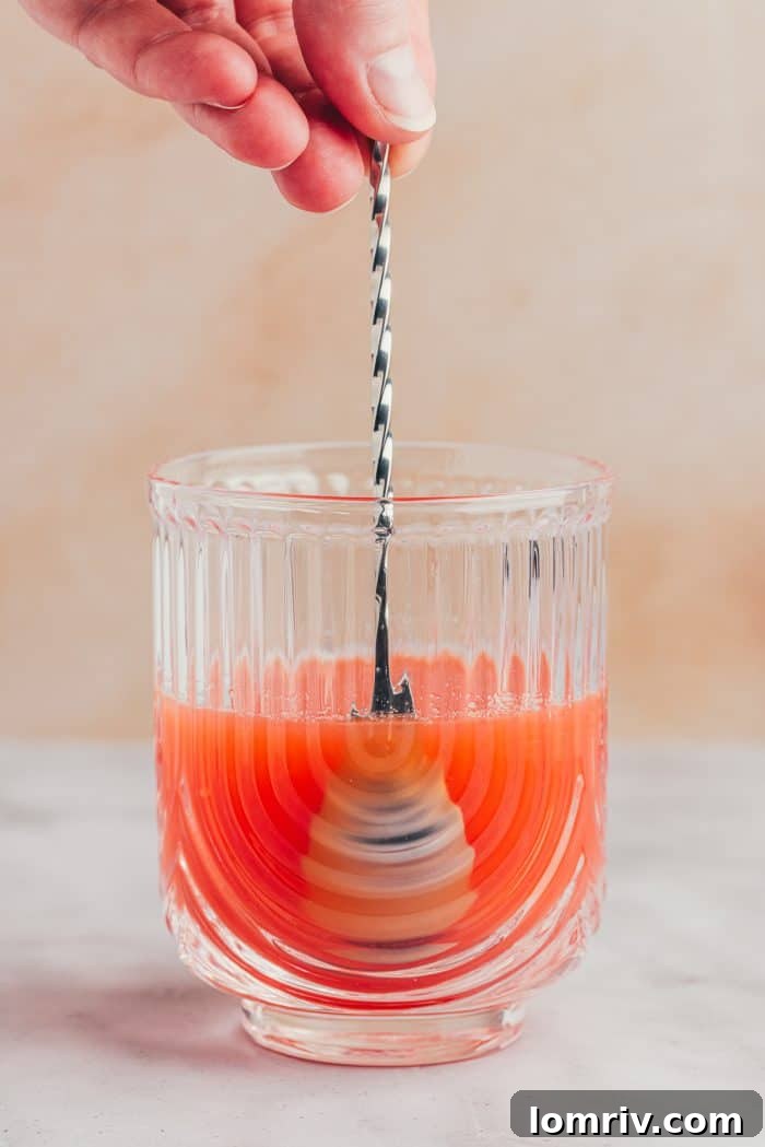 A small whisk stirring the blood orange, lime, and rosemary honey syrup mixture in a glass.