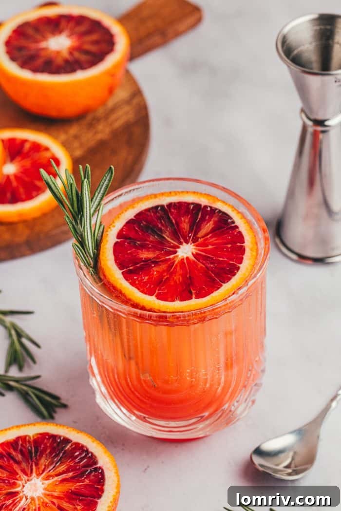 Three blood orange mocktails, each garnished with a blood orange slice and a rosemary sprig, on a rustic wooden surface.