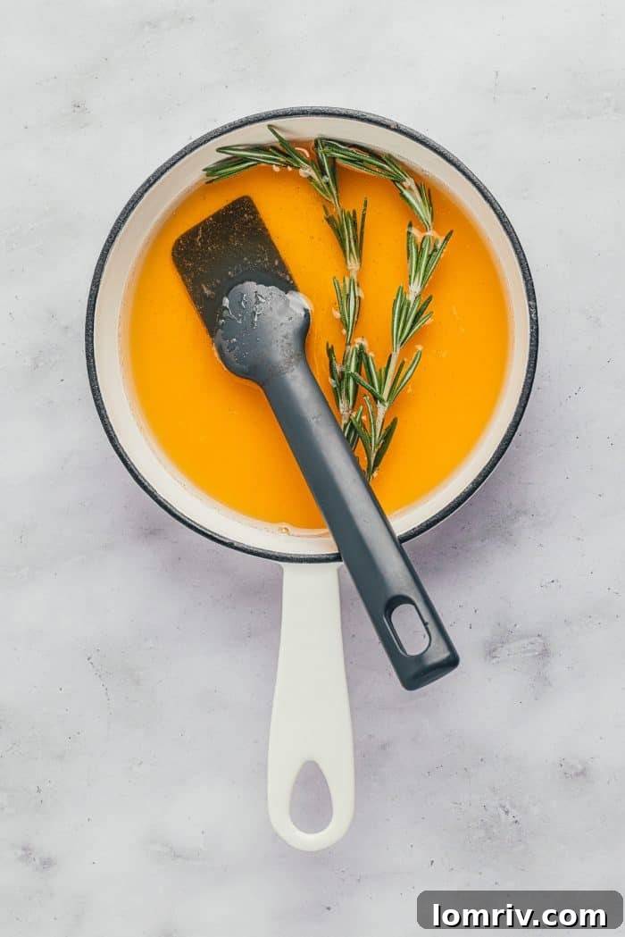 A small saucepan on a stovetop containing honey, water, and fresh rosemary sprigs, beginning to warm.
