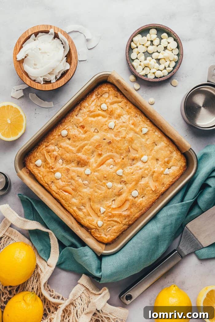 Freshly baked lemon blondies in a square baking tray, surrounded by lemons, coconut flakes, and white chocolate chips.