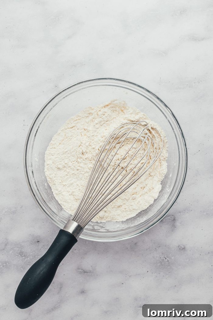 A clear glass bowl showcasing all-purpose flour being whisked with baking powder and salt.