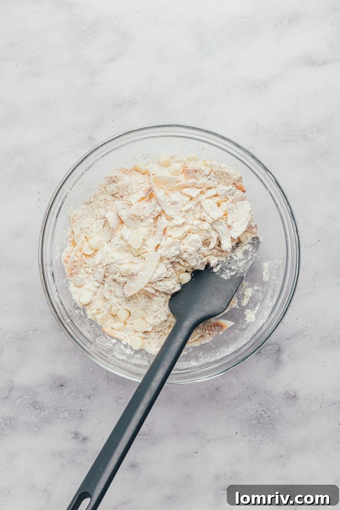 A glass bowl with flour, toasted coconut flakes, and white chocolate chips mixed together, with a spatula.