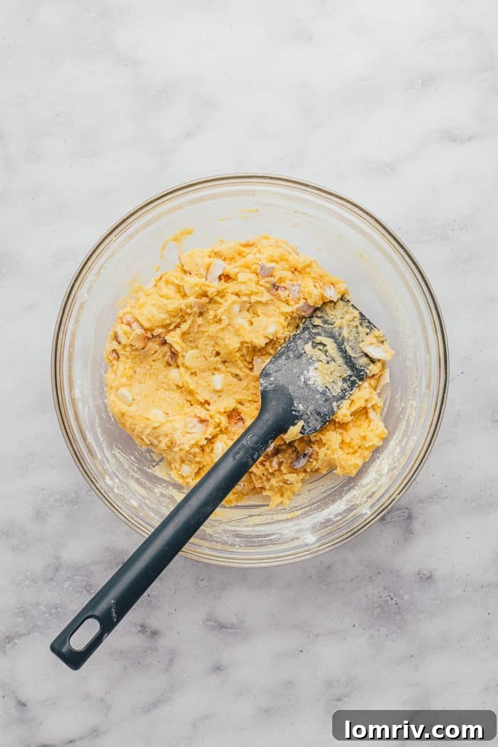 A glass bowl holding the thick, pale yellow lemon blondie batter with visible coconut and white chocolate chips, next to a rubber spatula.