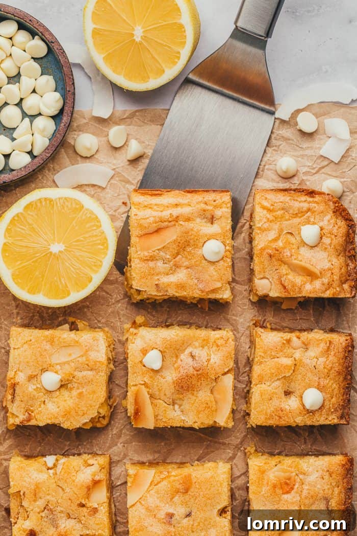 Lemon blondies being lifted from a baking pan with a spatula, with half a lemon and white chocolate chips in the background.