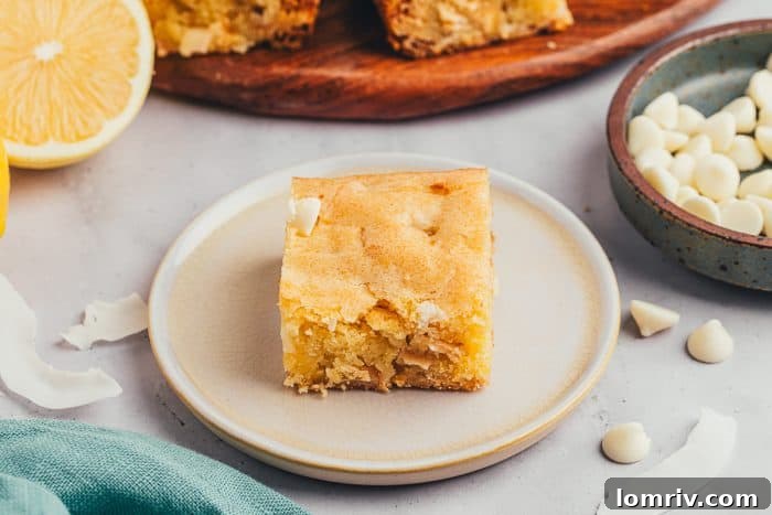 A single lemon blondie on a small plate, with a bowl of white chocolate chips in the background.