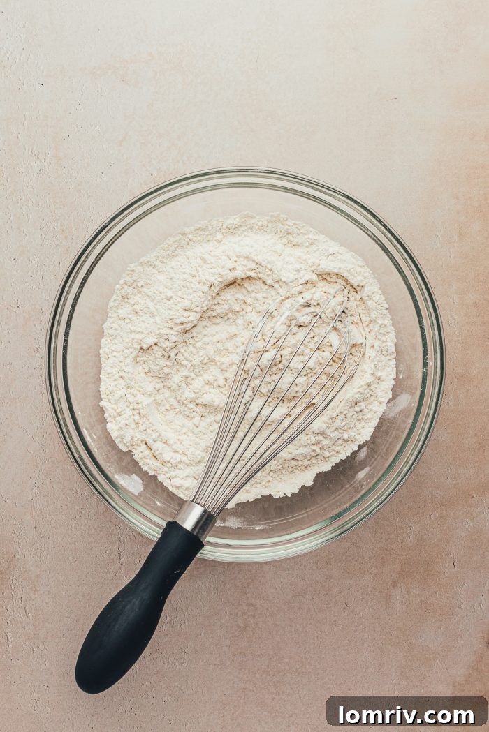 A separate glass bowl containing all-purpose flour, salt, and baking soda, with a whisk ready to combine them.