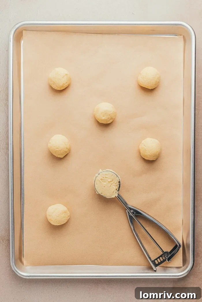 A cookie sheet lined with parchment paper, holding scoops of unbaked lemon sugar cookie dough, with a cookie scoop in hand.